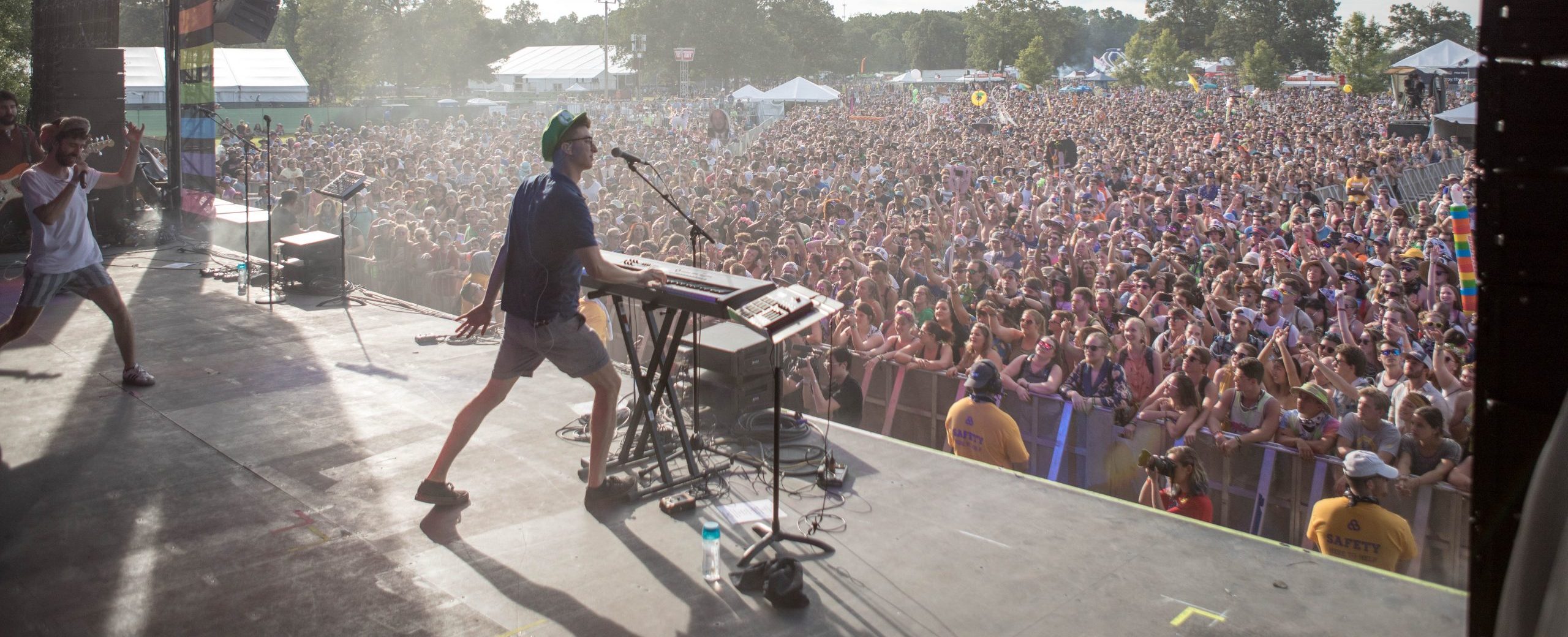 A musician plays keyboard and sings on stage at an outdoor music festival, facing a large cheering crowd. Security staff stand by the barricade, and tents and trees are visible in the background.