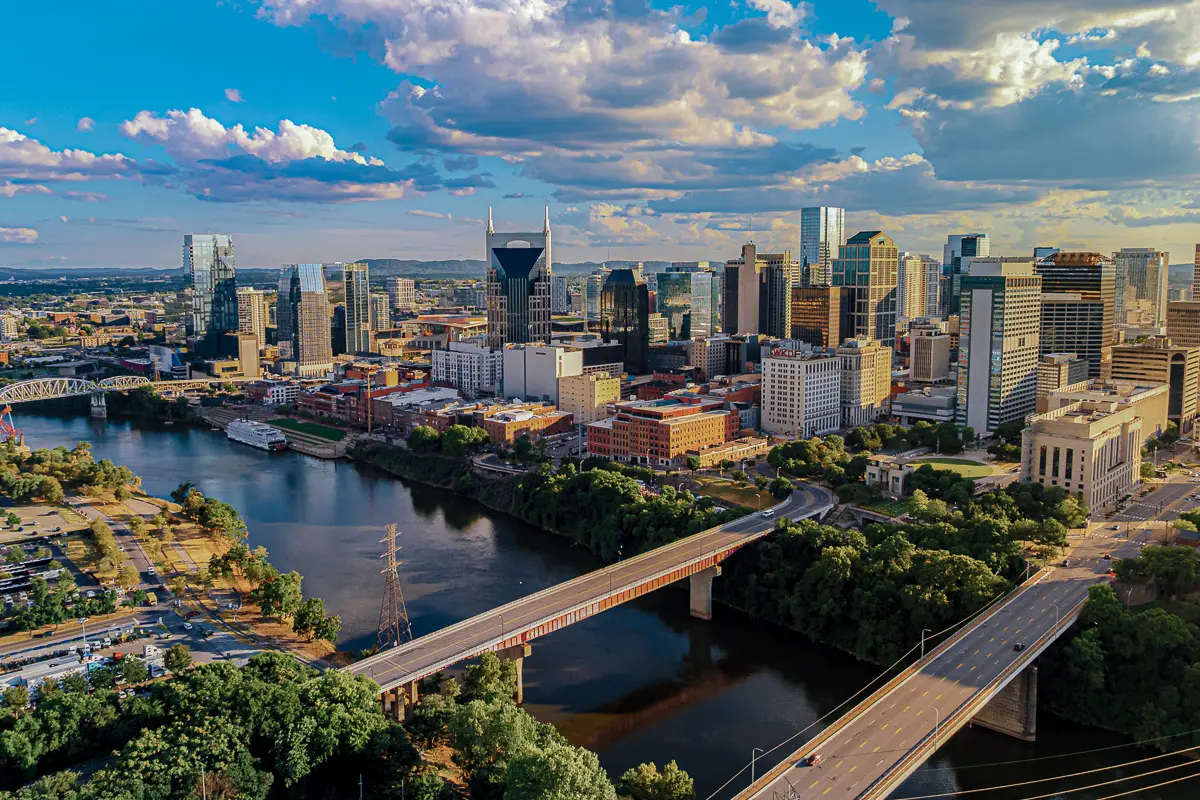 Aerial view of downtown Nashville, Tennessee, featuring the Cumberland River, bridges, and modern city skyline under a partly cloudy sky.