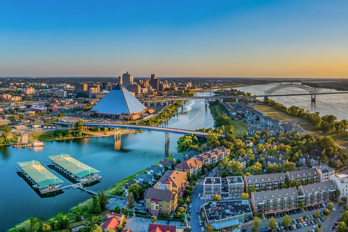 Aerial view of downtown Memphis, Tennessee at sunset, featuring the Mississippi River, a large glass pyramid, bridges, and clusters of buildings and greenery along the waterfront.