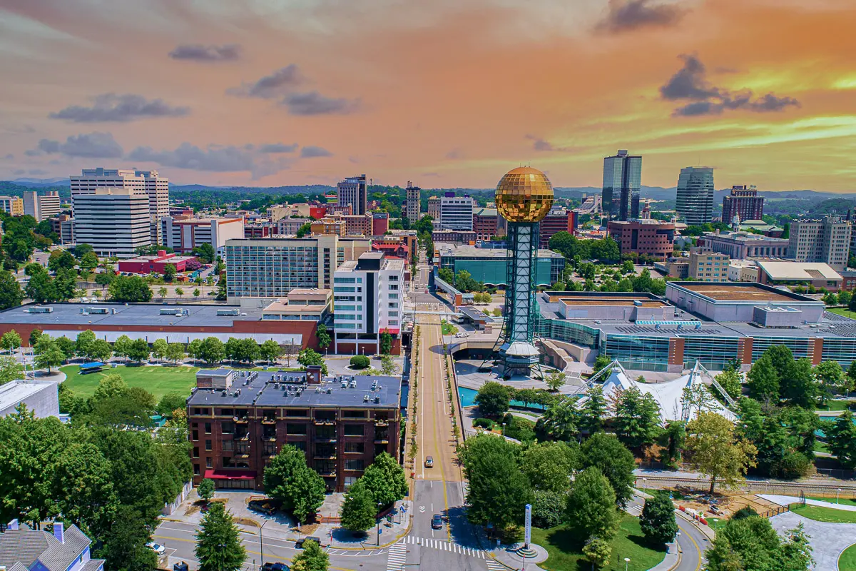 Aerial view of downtown Knoxville, Tennessee, featuring the Sunsphere, modern and historic buildings, green spaces, and a vibrant sunset sky with orange and purple hues.