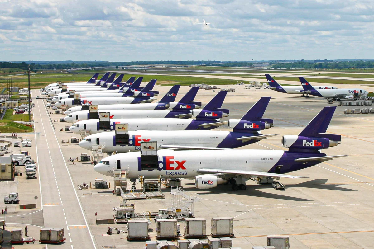 A row of FedEx Express cargo airplanes is parked on the tarmac at an airport, with ground service vehicles nearby and green fields in the background under a partly cloudy sky.