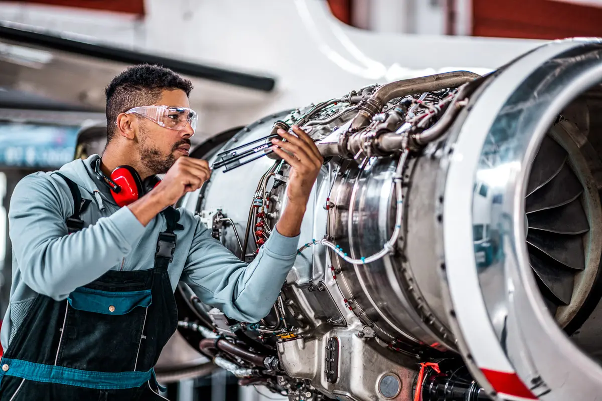 A mechanic wearing safety glasses and headphones works on a jet engine, using tools to adjust its components. The engine’s intricate wiring and parts are visible as he focuses on the task.