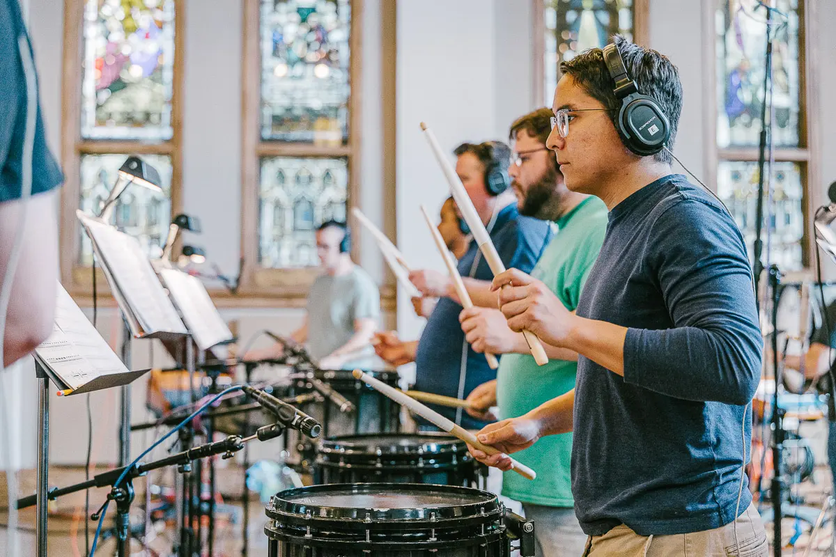 Several people wearing headphones play drums with drumsticks inside a bright room with stained-glass windows. Sheet music stands and microphones are visible around them.