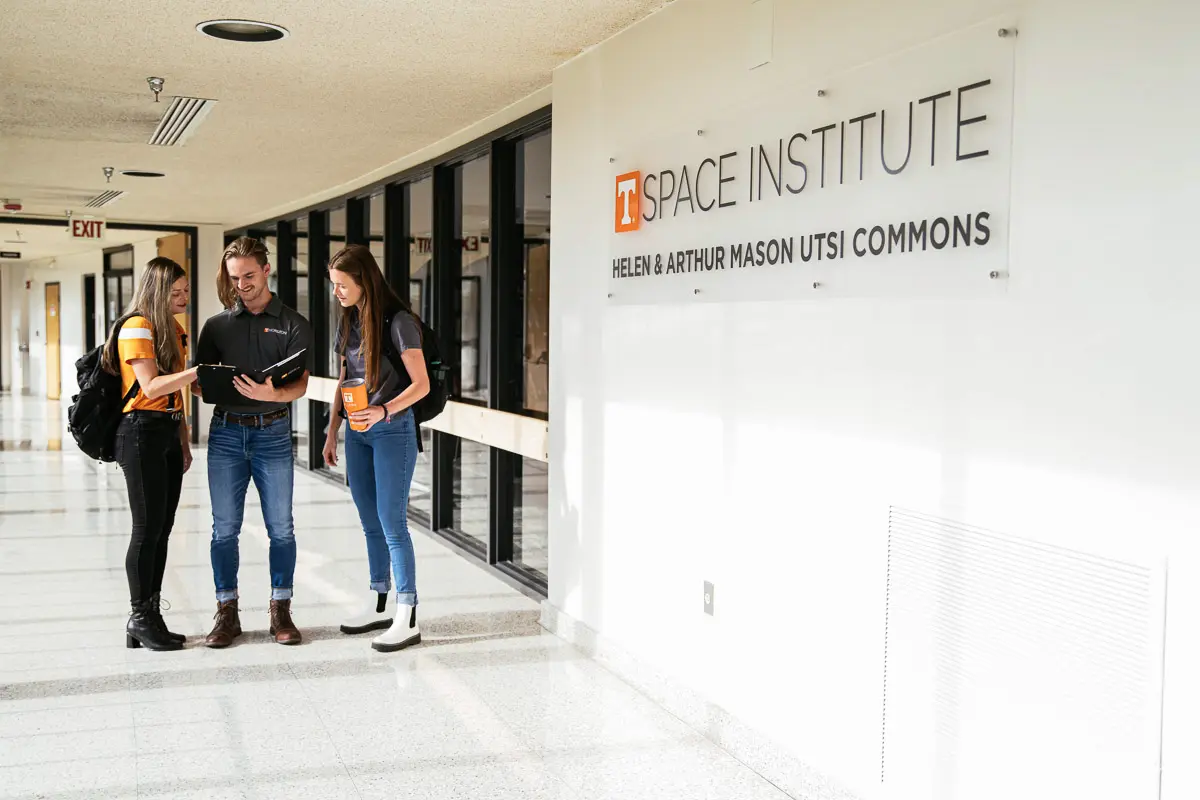 Three people stand in a bright hallway near a sign that reads “Space Institute, Helen & Arthur Mason UTSI Commons,” talking and looking at a tablet. Two are students with backpacks; one appears to be a staff member.