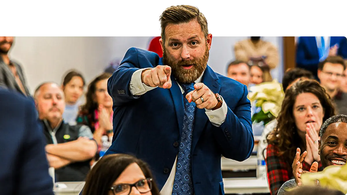 A bearded man in a blue suit enthusiastically points toward the camera in a crowded room, surrounded by people sitting at tables, some smiling and clapping.