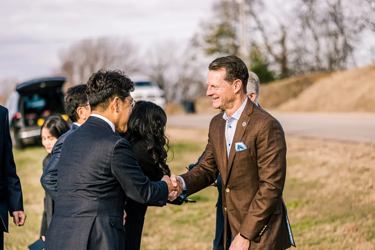 A group of people in business attire greet each other outdoors. Two men in the foreground shake hands and smile, with cars and dry grass in the background.