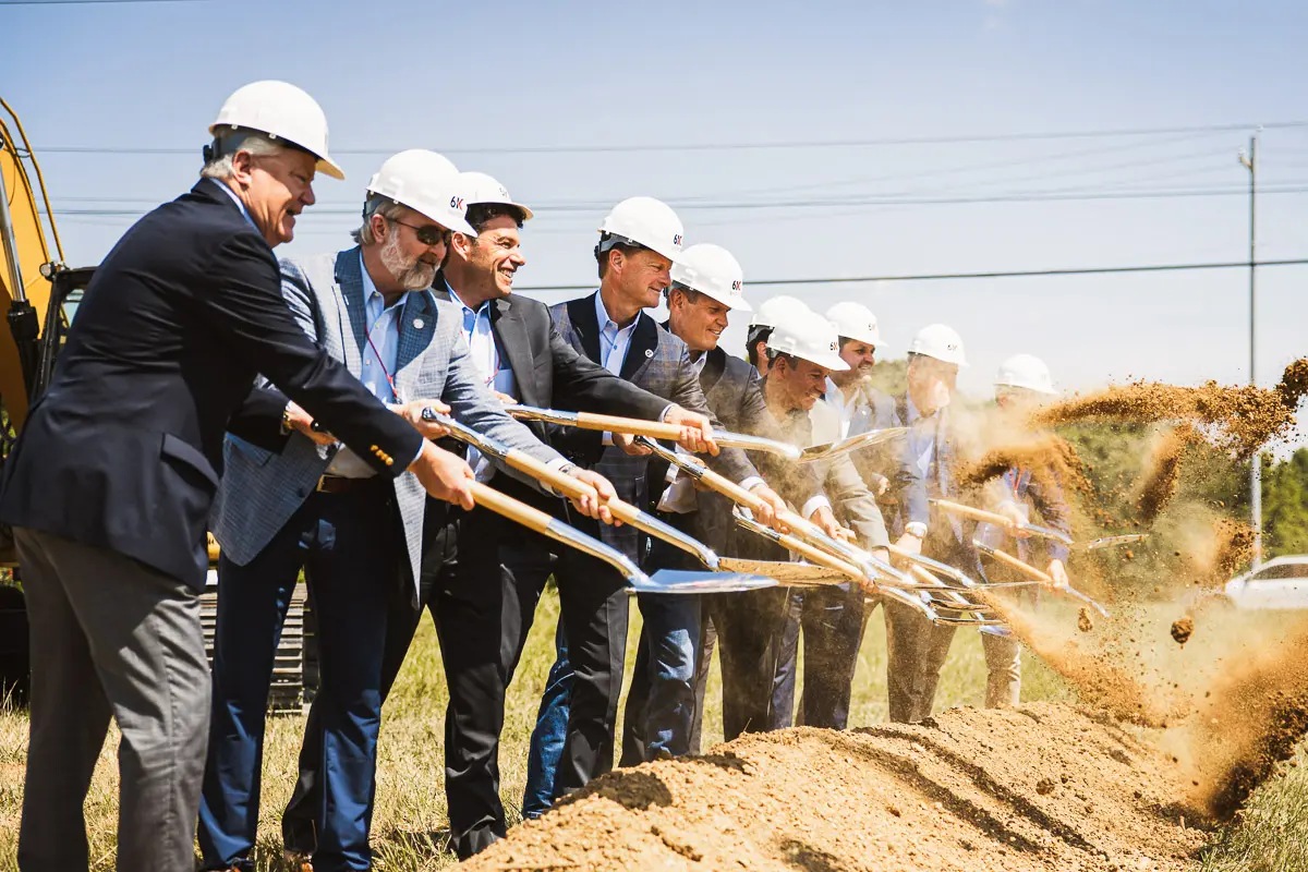 A group of people in business attire and white hard hats use shovels to break ground at a construction site, tossing dirt into the air during a sunny outdoor ceremony.