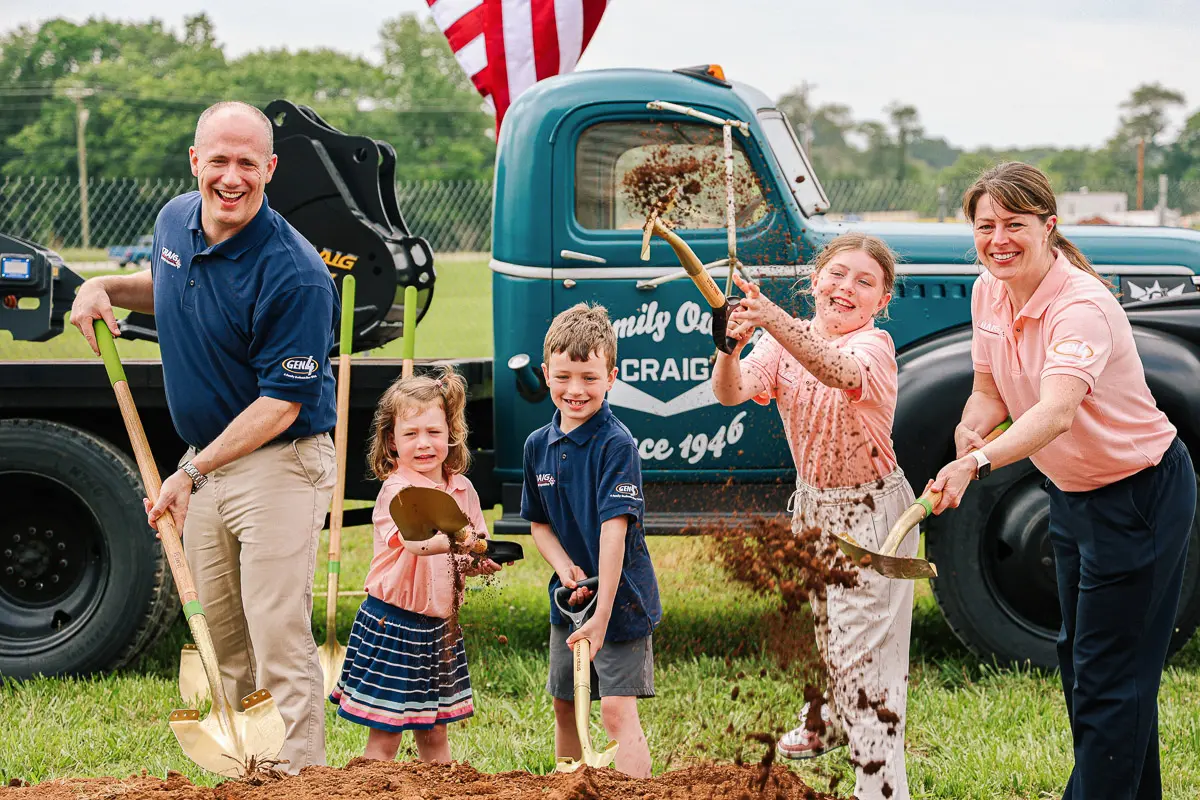 A smiling family of five, including three children, uses shovels to toss dirt at a groundbreaking ceremony outdoors, with an American flag and a vintage truck in the background.