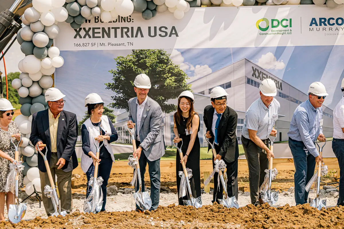 A group of people in hard hats and business attire smile and pose with shovels at a groundbreaking ceremony in front of a sign that reads "XXENTRIA USA." Balloons and a building rendering are visible in the background.