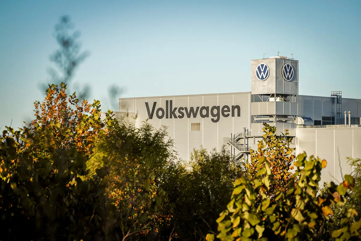 A Volkswagen factory building with the company name and logo, partially obscured by green and yellow trees under a clear blue sky.