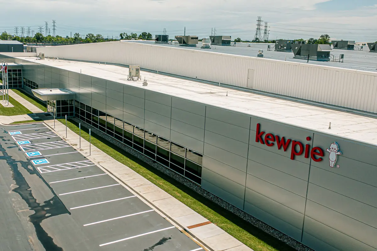 A large, modern industrial building with "kewpie" and the Kewpie doll logo on the side, next to an empty parking lot with several accessible parking spaces marked.