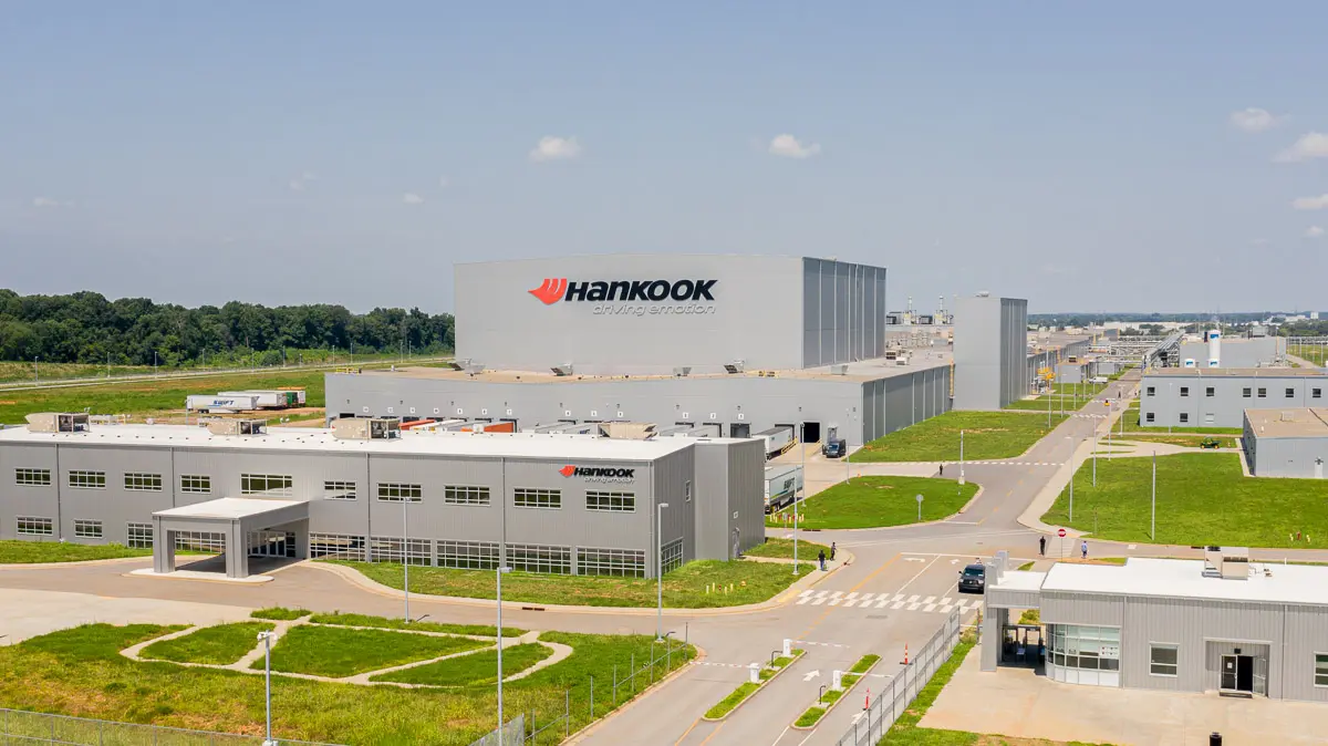 A large, modern Hankook facility with multiple gray buildings, green lawns, and paved roads, seen under a clear blue sky. The main building displays the Hankook logo and the slogan "driving emotion.