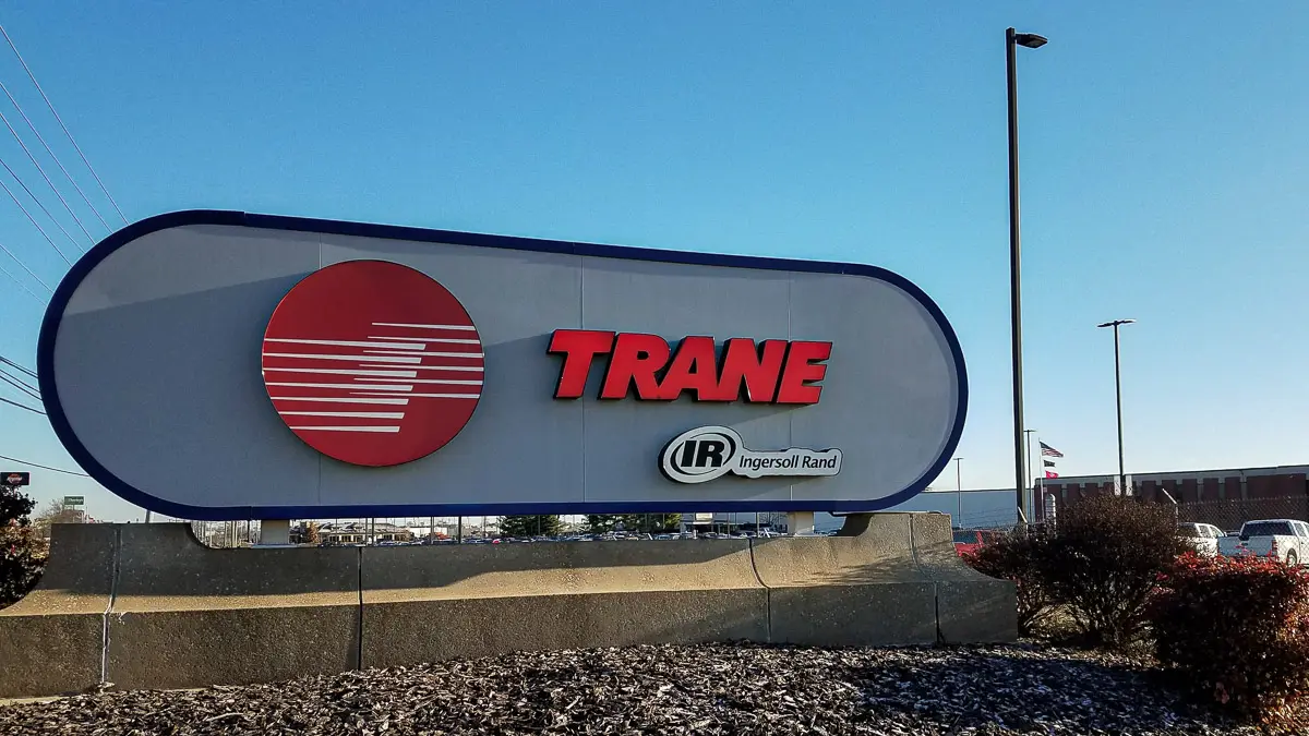 Large outdoor sign with a red and white circular logo, the word "TRANE" in red letters, and "IR Ingersoll Rand" below, set against a clear blue sky and industrial buildings in the background.