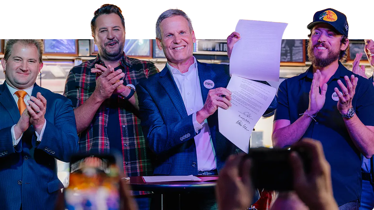 Four men stand together on a stage. The man in the center, smiling, holds up and displays a signed document while the others clap and smile. People in the foreground take photos of the moment.