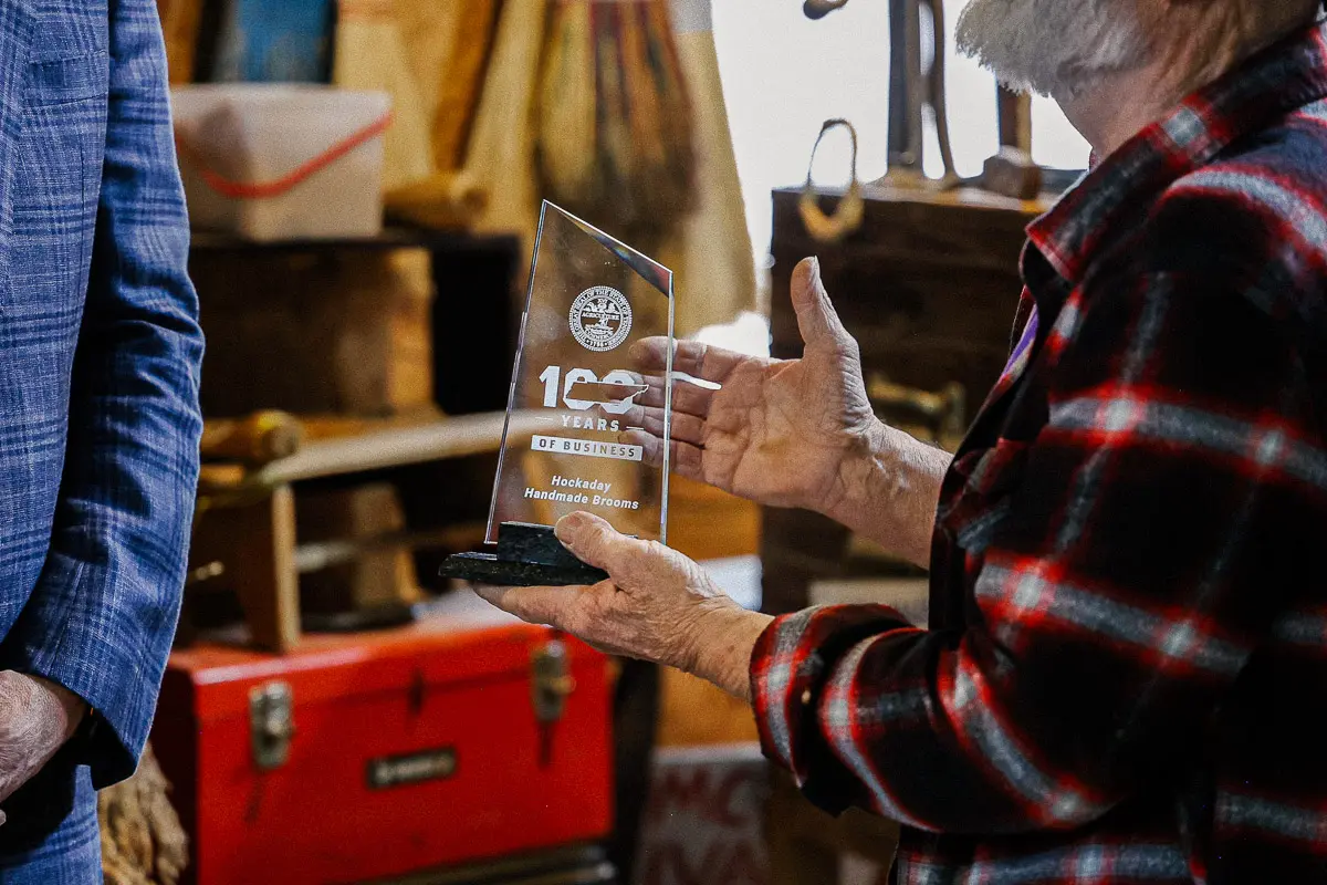 A person in a red plaid shirt holds a glass award that reads "100 Years of Business" with an emblem above it. Another person stands nearby. The background shows rustic wooden crates and a red toolbox.