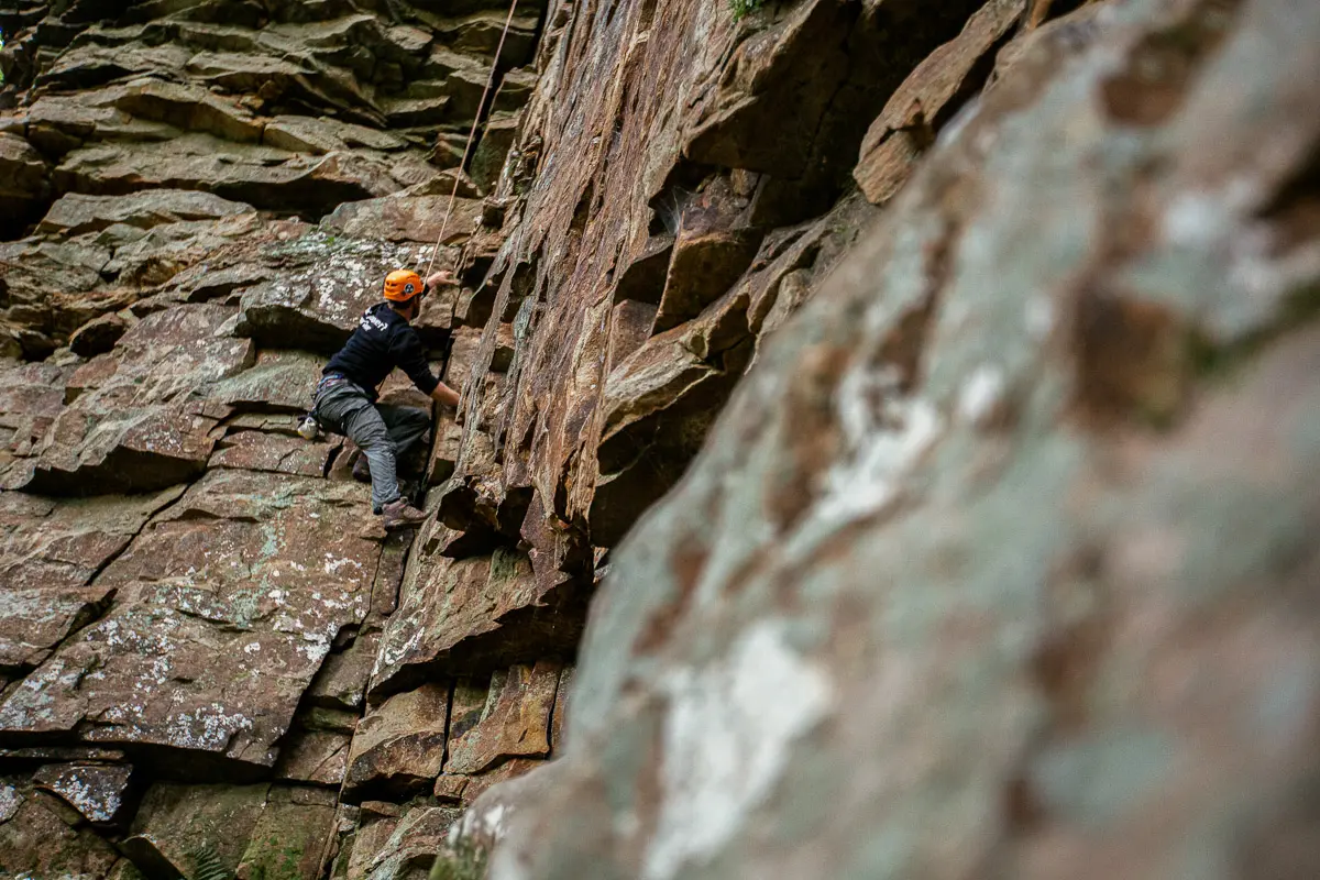 A person wearing an orange helmet and climbing gear scales a steep, rugged rock face outdoors, using both hands and feet to grip the uneven surface.