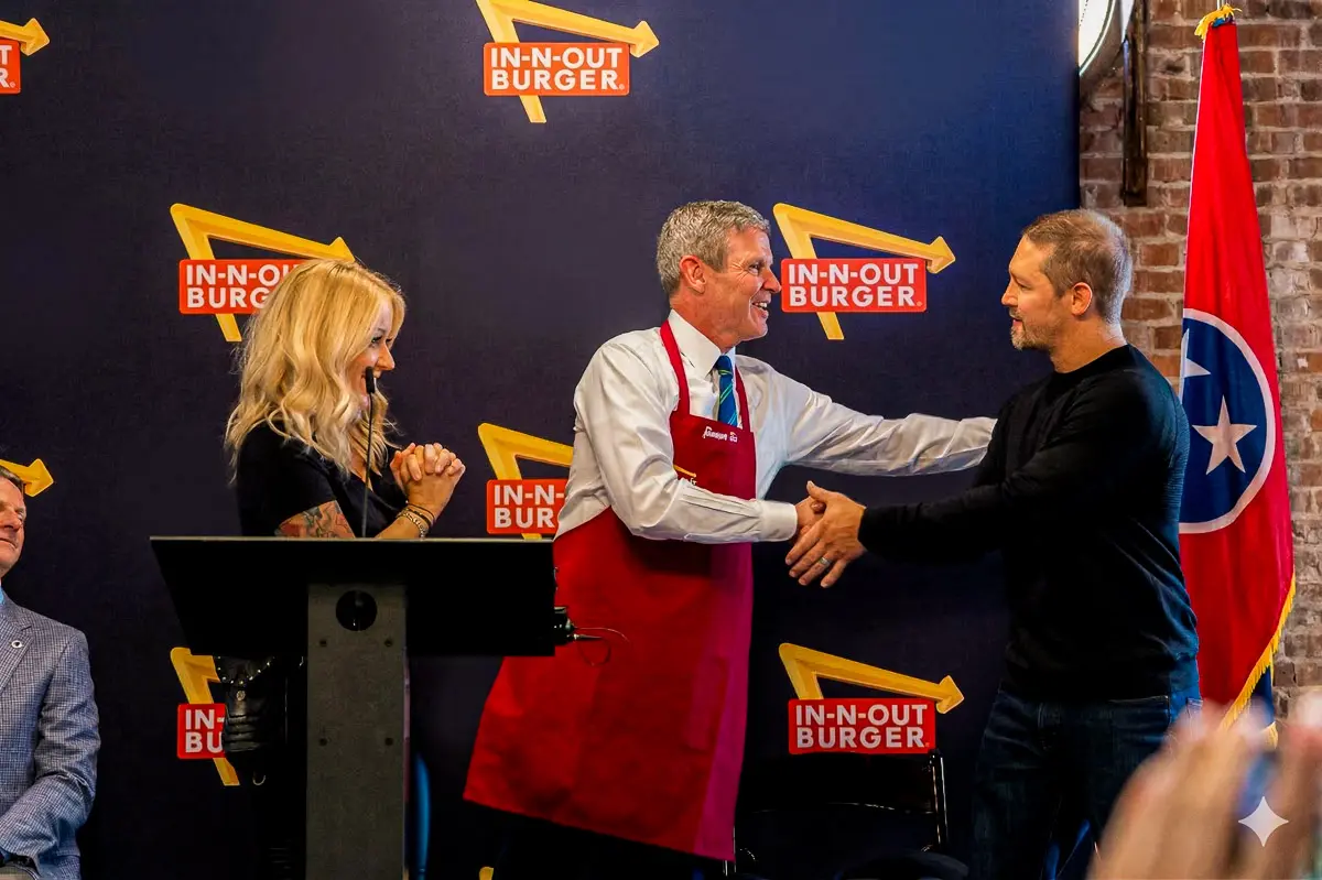 A man in a red apron shakes hands with another man in front of a backdrop featuring multiple In-N-Out Burger logos, while a woman smiles and stands at a podium. A Tennessee state flag is visible on the right.