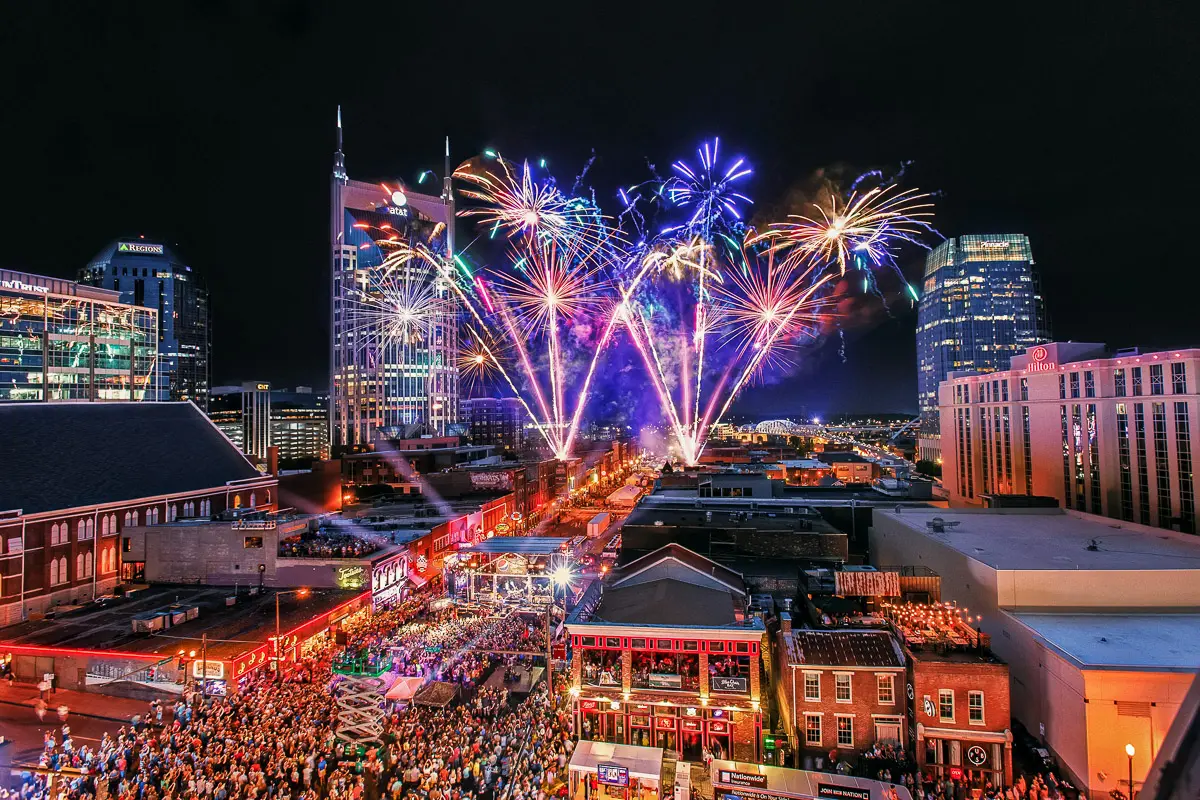 A vibrant cityscape at night shows colorful fireworks bursting above tall buildings and a lively, crowded street filled with people, illuminated by neon lights from bars and restaurants.