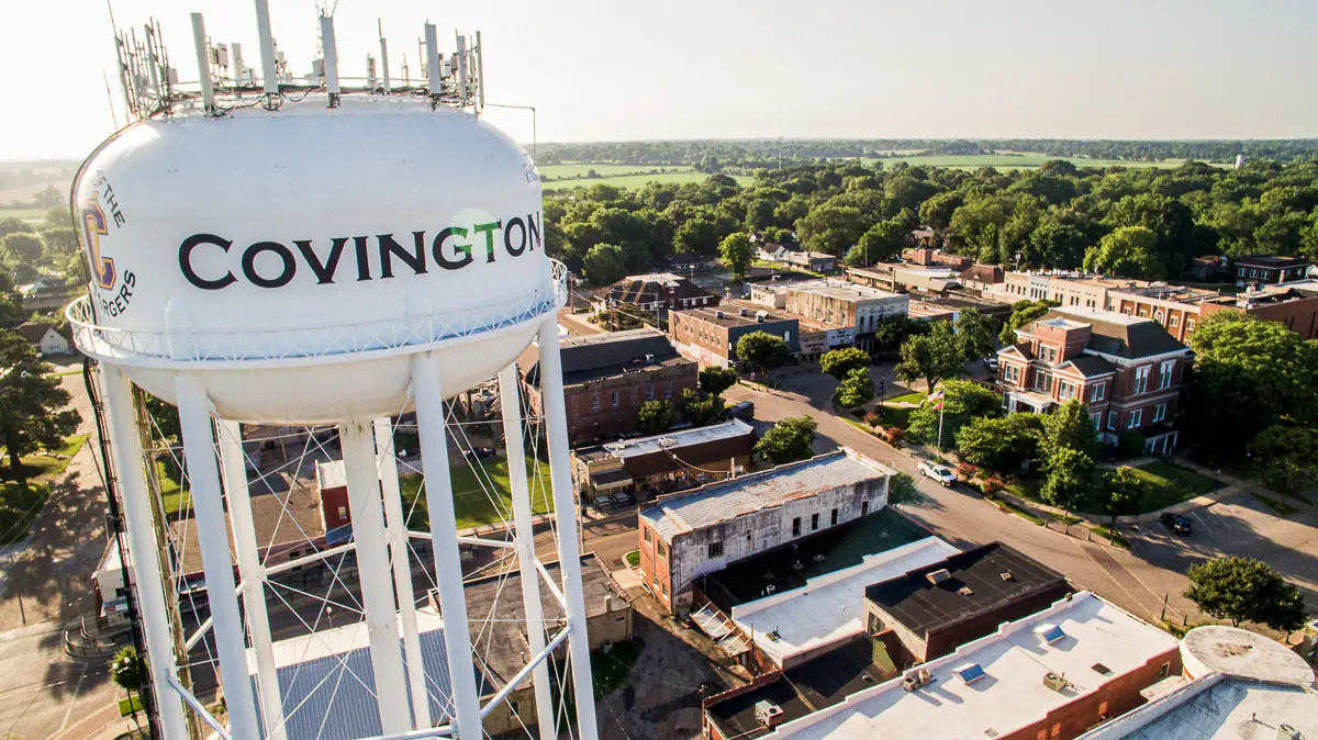 A large white water tower labeled "Covington" stands above a small town with tree-lined streets, brick buildings, and green fields stretching into the distance under a clear sky.