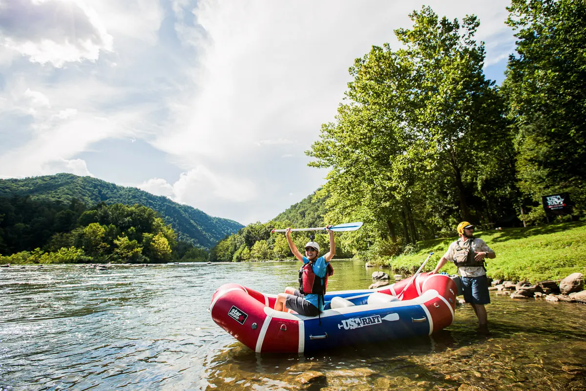 Two people with a red inflatable raft stand in a scenic river surrounded by green hills and trees; one person raises a paddle overhead, celebrating, while the other stands nearby in the shallow water.