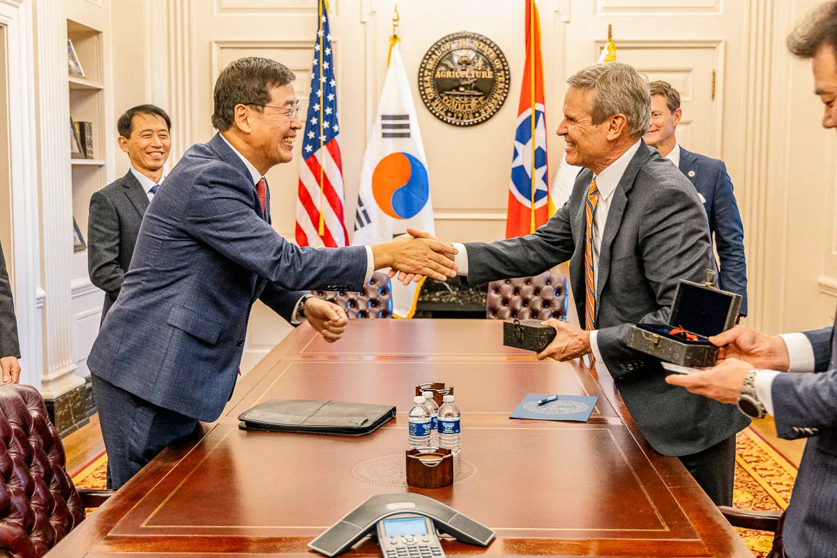 Two men in suits shake hands across a conference table, smiling. Behind them are South Korean and Tennessee state flags. Other people in suits stand nearby, and gift boxes are exchanged.