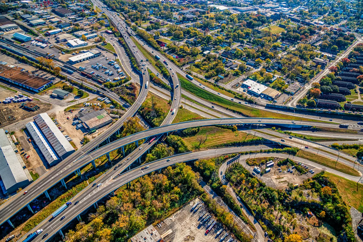Aerial view of a sprawling highway interchange with multiple overpasses, surrounded by trees, industrial buildings, neighborhoods, and green spaces on a sunny day.