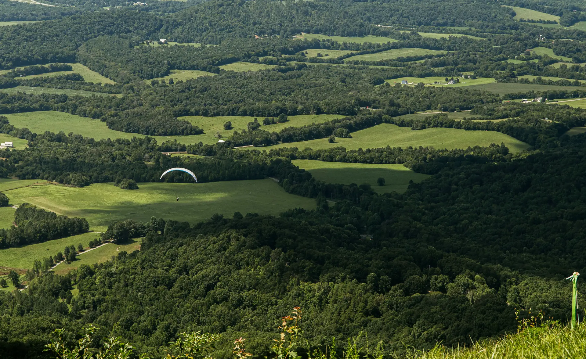 A paraglider glides over a patchwork of green fields and dense forests with rolling hills under a clear sky, seen from a high vantage point.