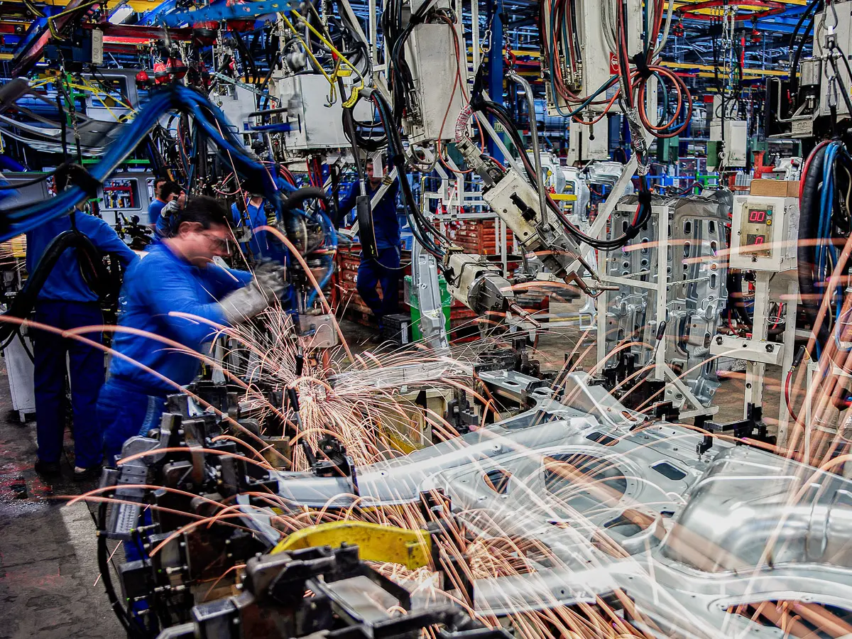 Factory workers operate machinery as sparks fly from robotic arms welding metal car parts on an automotive assembly line. The scene is busy, with various cables and equipment surrounding the workspace.