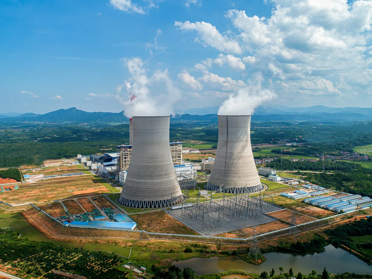 Aerial view of a power plant with two large cooling towers releasing steam into the sky, surrounded by green fields, buildings, and distant mountains under a partly cloudy sky.