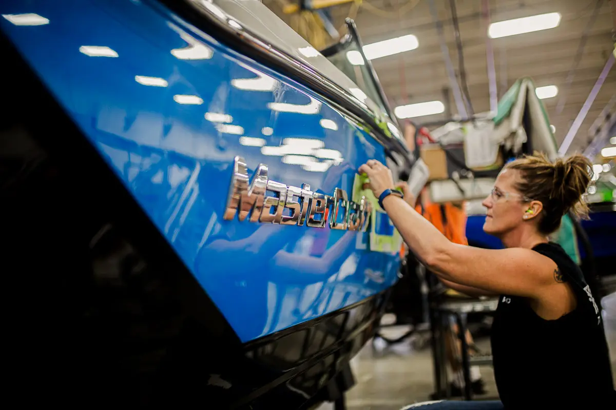 A woman wearing ear protection works on the side of a blue MasterCraft boat in an indoor workshop, carefully applying or inspecting details on the boat’s exterior.