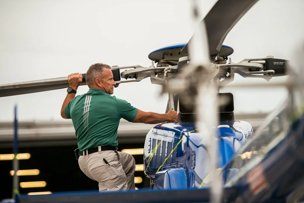 A man in a green shirt and khaki pants inspects or repairs the rotor blades of a blue helicopter, with part of the helicopter visible in the foreground.