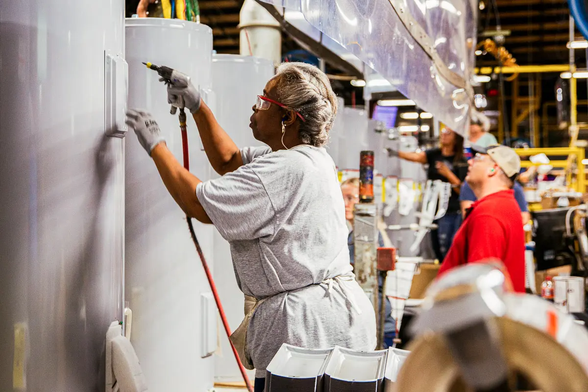 A woman wearing safety glasses and gloves works on a large industrial tank in a busy factory, using a pneumatic tool. Other workers are seen in the background, focused on their tasks.