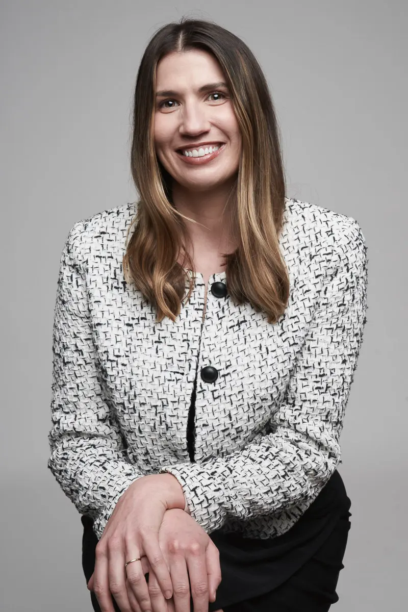 A woman with long, straight brown hair is smiling at the camera. She is wearing a white textured jacket with black patterns and buttons, and has her hands crossed in front of her. The background is plain light gray.