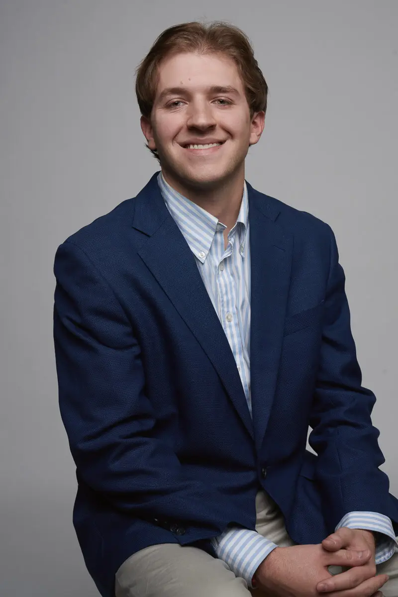 A young man with light brown hair, wearing a blue blazer, light blue striped shirt, and khaki pants, smiles while sitting against a plain gray background.