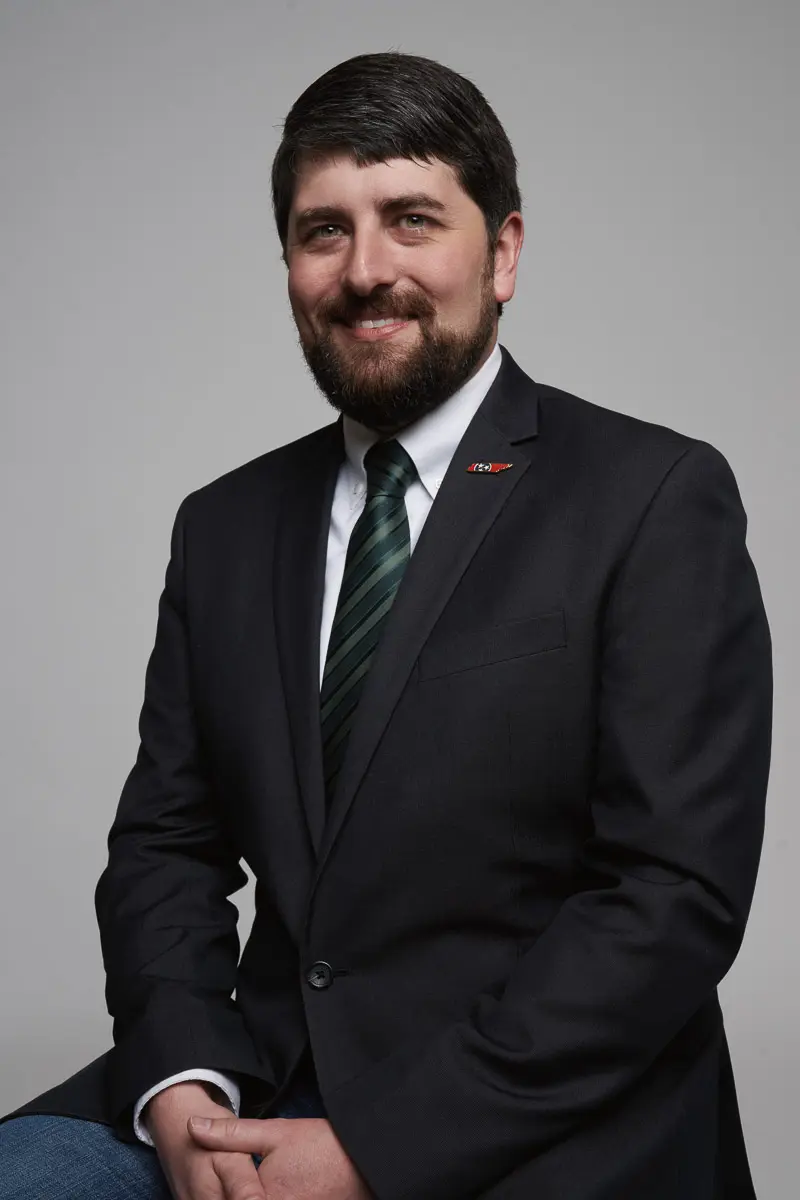 A man with dark hair and a beard, wearing a dark suit, white shirt, and green striped tie, sits and smiles at the camera against a plain gray background.