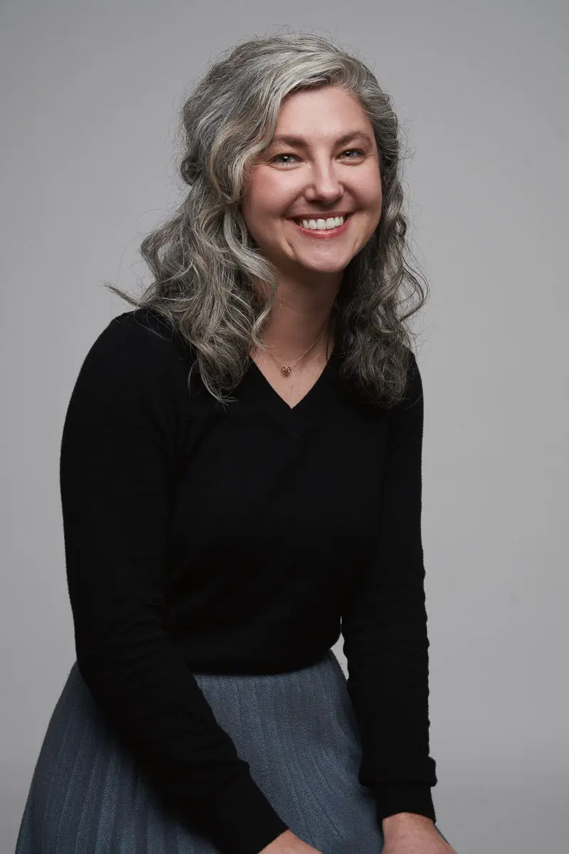 A woman with wavy gray hair, wearing a black long-sleeve top and a blue-gray skirt, smiles while sitting against a plain gray background.
