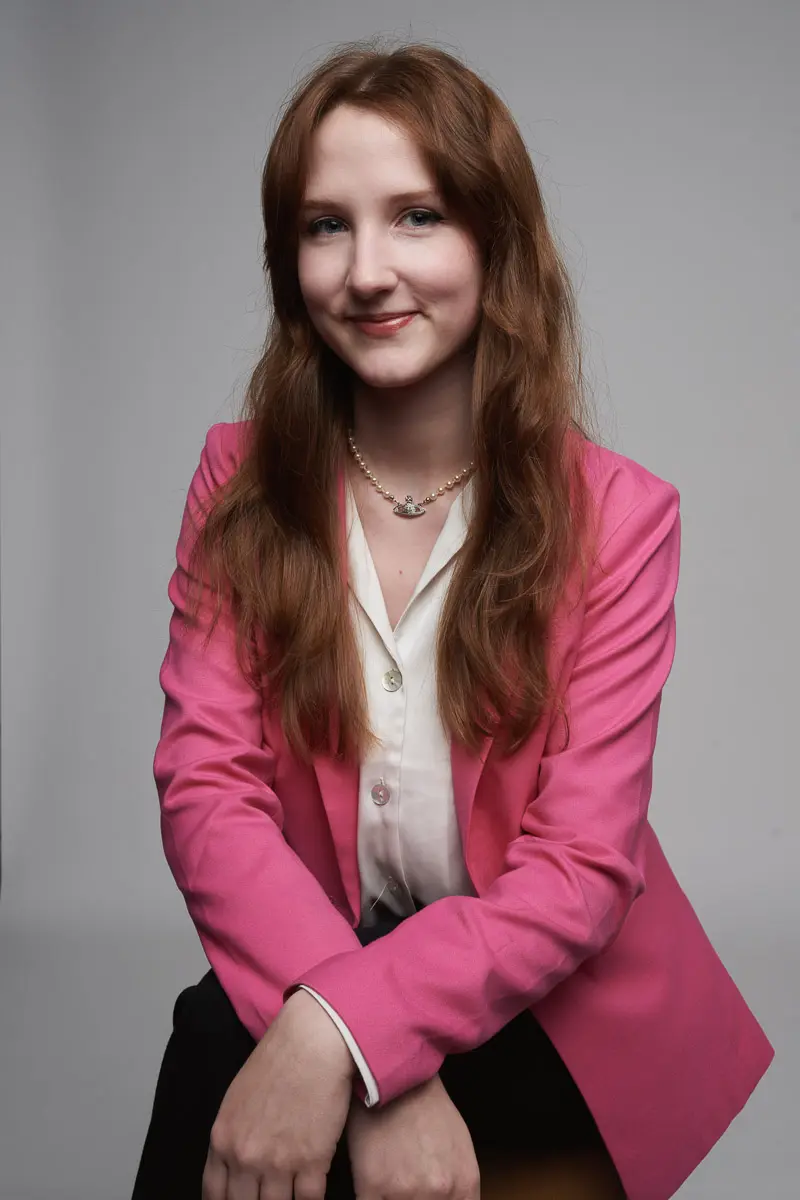 A young woman with long, wavy brown hair sits smiling. She wears a bright pink blazer over a white blouse and a delicate pearl necklace. The background is plain gray, and she has her hands resting in her lap.