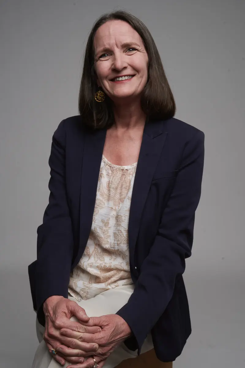 A middle-aged woman with straight brown hair sits on a stool against a plain background, smiling warmly. She wears a navy blazer over a patterned blouse and cream pants, with her hands resting on her knee.
