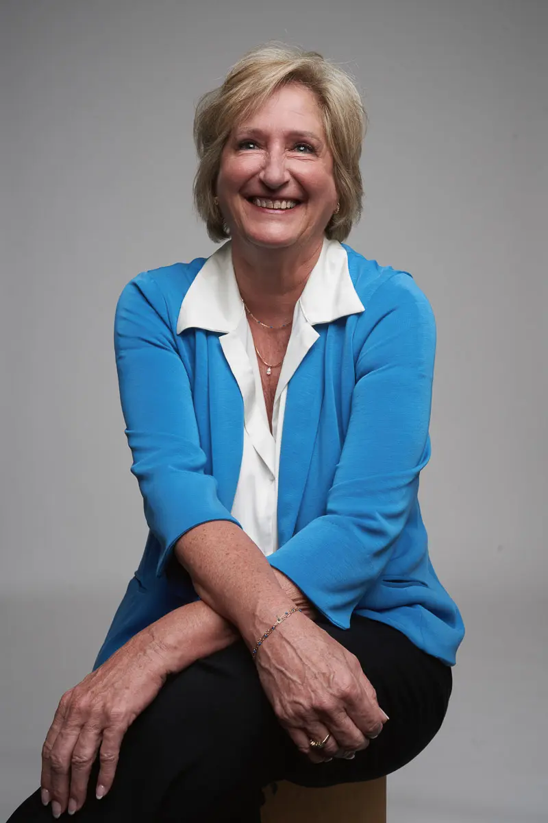 A smiling older woman with short blonde hair wearing a blue cardigan over a white blouse and black pants, sits on a stool with her arms resting on her knee against a plain gray background.