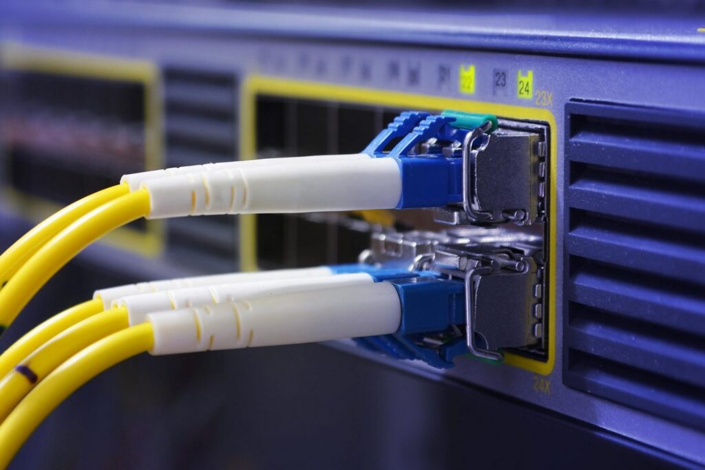 Close-up of yellow fiber optic cables plugged into network equipment ports, with blue connectors and a blurred background of a data center device.
