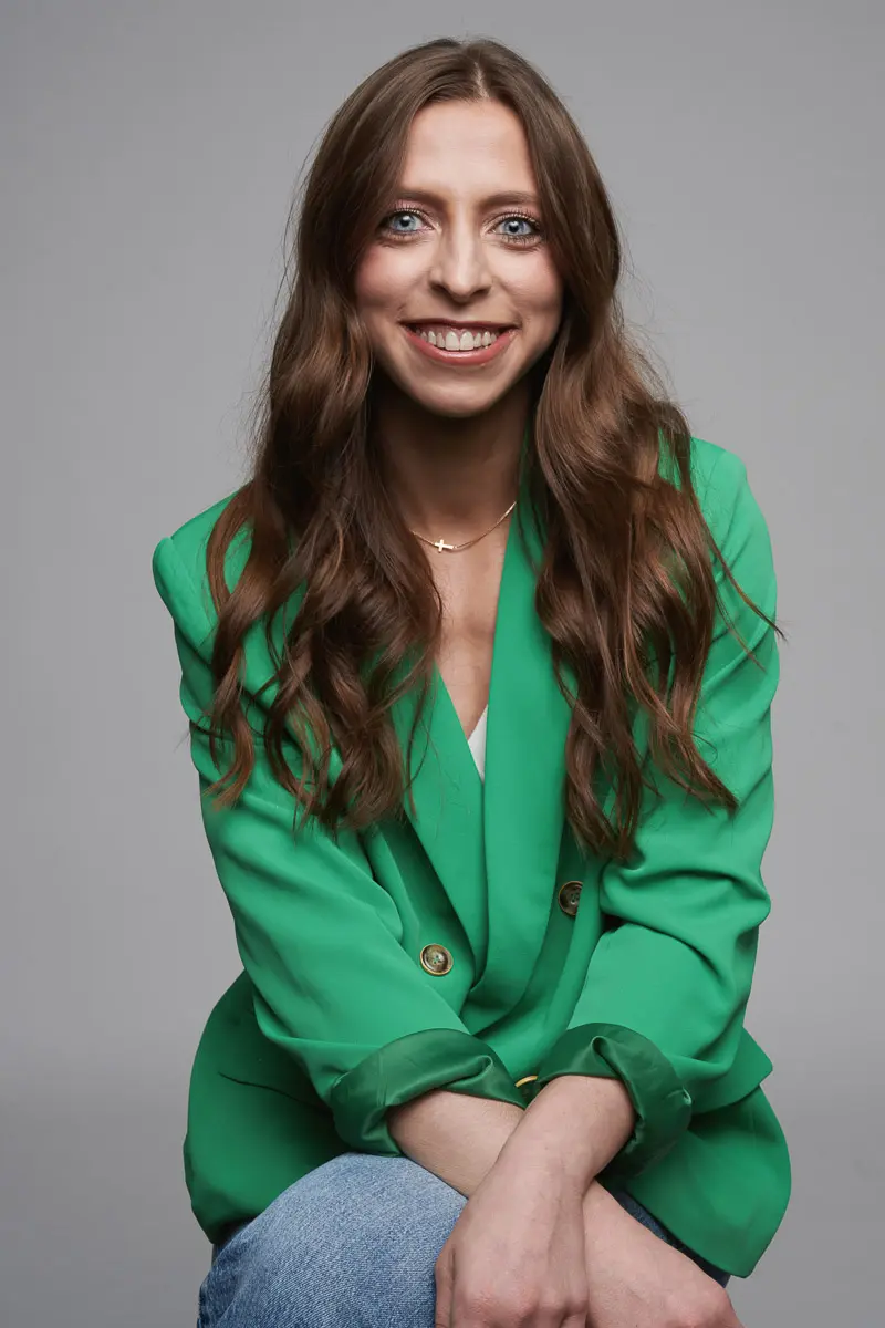 A woman with long brown hair wearing a bright green blazer and blue jeans sits smiling against a plain gray background.