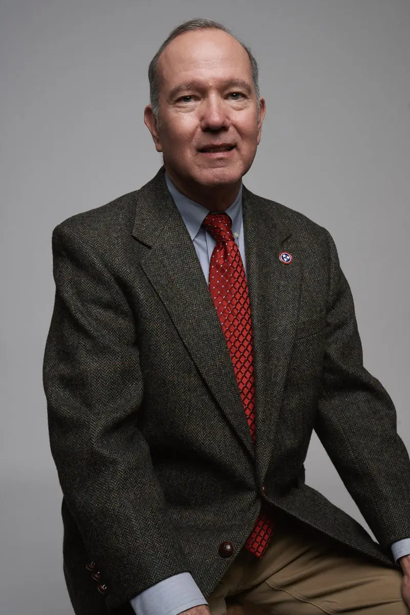 A middle-aged man with short gray hair wearing a dark textured blazer, light blue shirt, and red patterned tie, sitting and facing forward against a plain gray background.