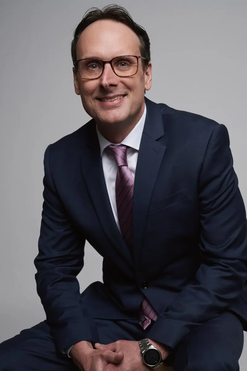 A man in a navy suit, white shirt, and pink striped tie sits smiling at the camera. He wears glasses and a watch, and has short, dark hair. The background is plain and light gray.