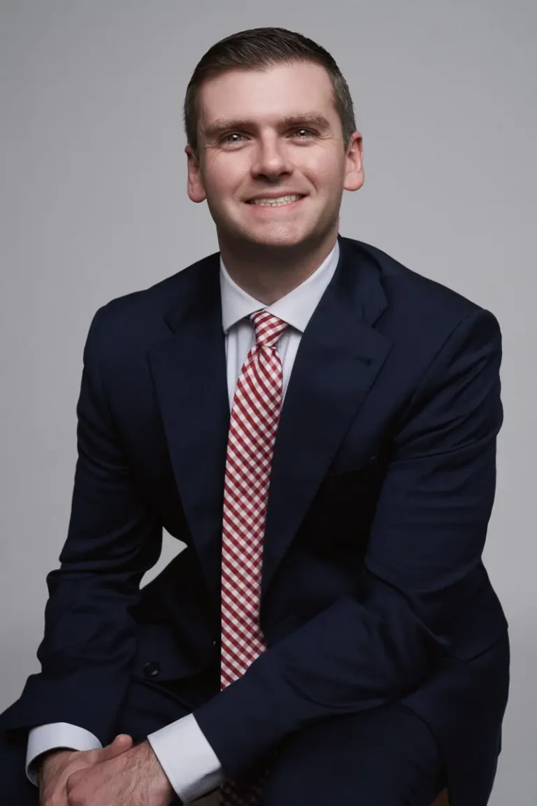 A man in a navy blue suit and red-and-white striped tie sits on a stool, smiling at the camera against a plain gray background.