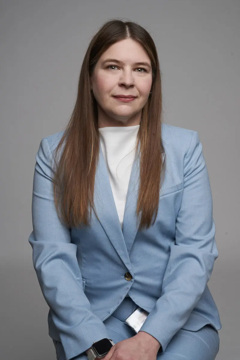 A woman with long brown hair wearing a light blue suit and white top sits against a plain gray background, facing forward with a neutral expression. She has a smartwatch on her left wrist.