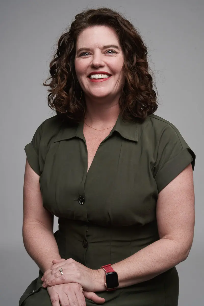 A woman with curly brown hair wearing an olive green short-sleeve dress and a smartwatch sits smiling against a plain gray background.