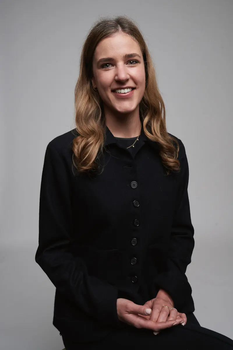 A woman with long, wavy brown hair, wearing a black buttoned-up shirt, sits and smiles at the camera against a plain, light gray background. Her hands are gently folded on her lap.
