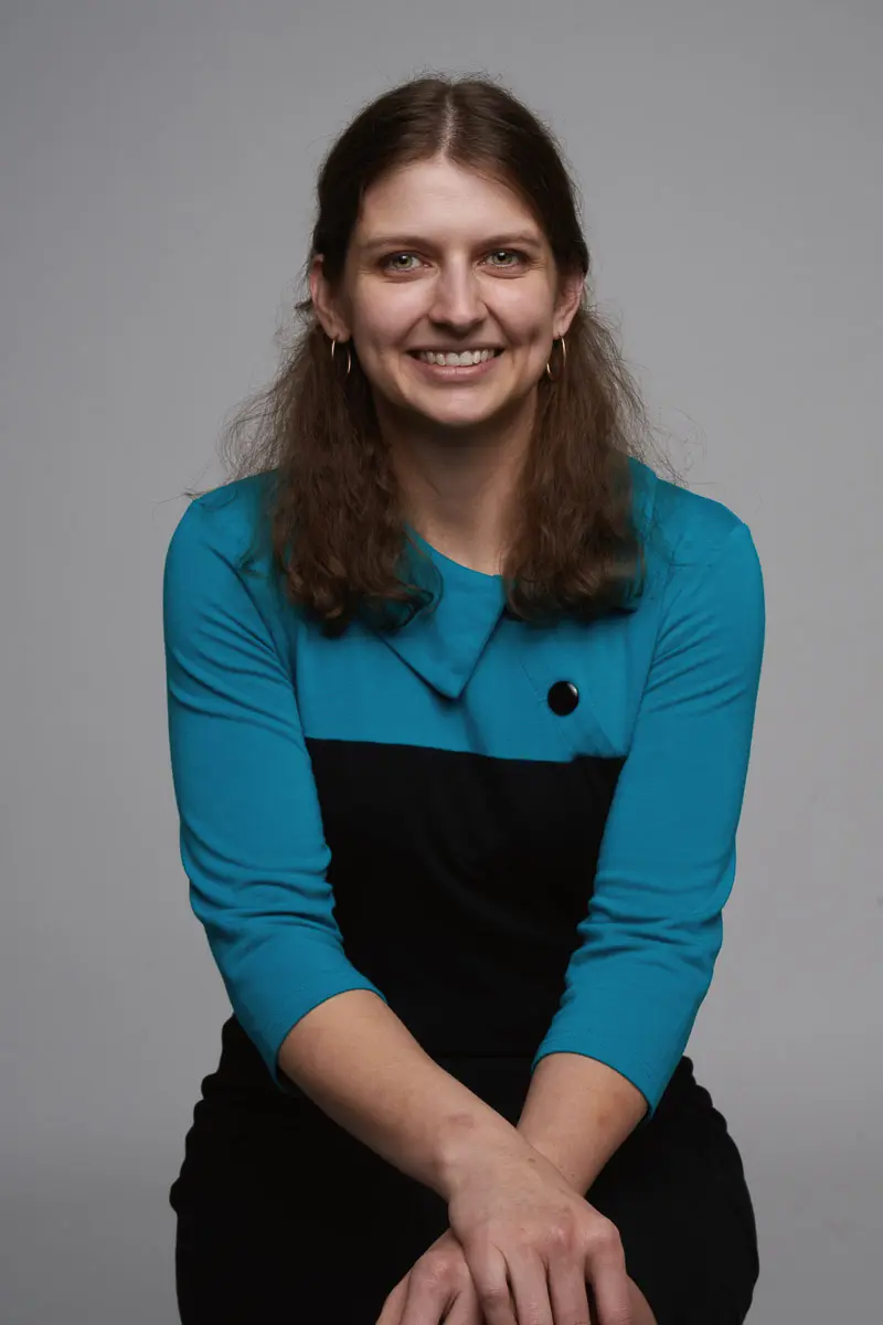 A woman with long brown hair, wearing a teal and black dress with a button detail, sits against a plain gray background, smiling at the camera with her hands resting in her lap.