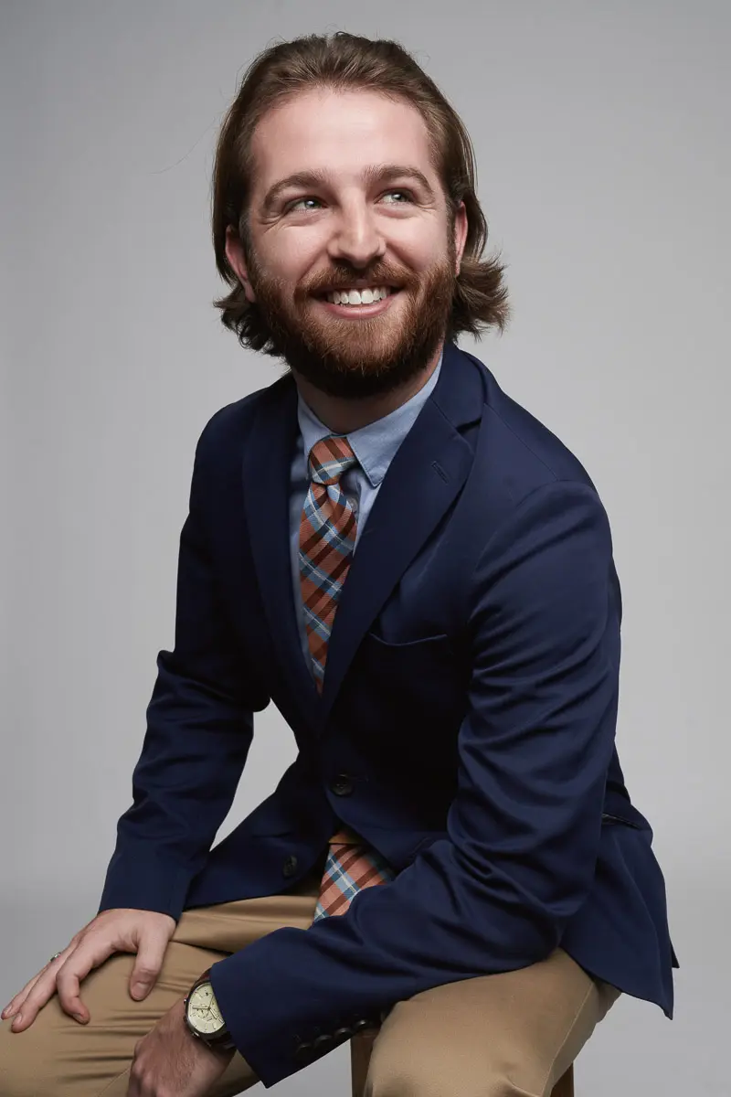 A man with shoulder-length brown hair and a beard smiles while sitting on a stool. He is wearing a navy blazer, light blue shirt, striped tie, tan pants, and a watch, with a neutral gray background.