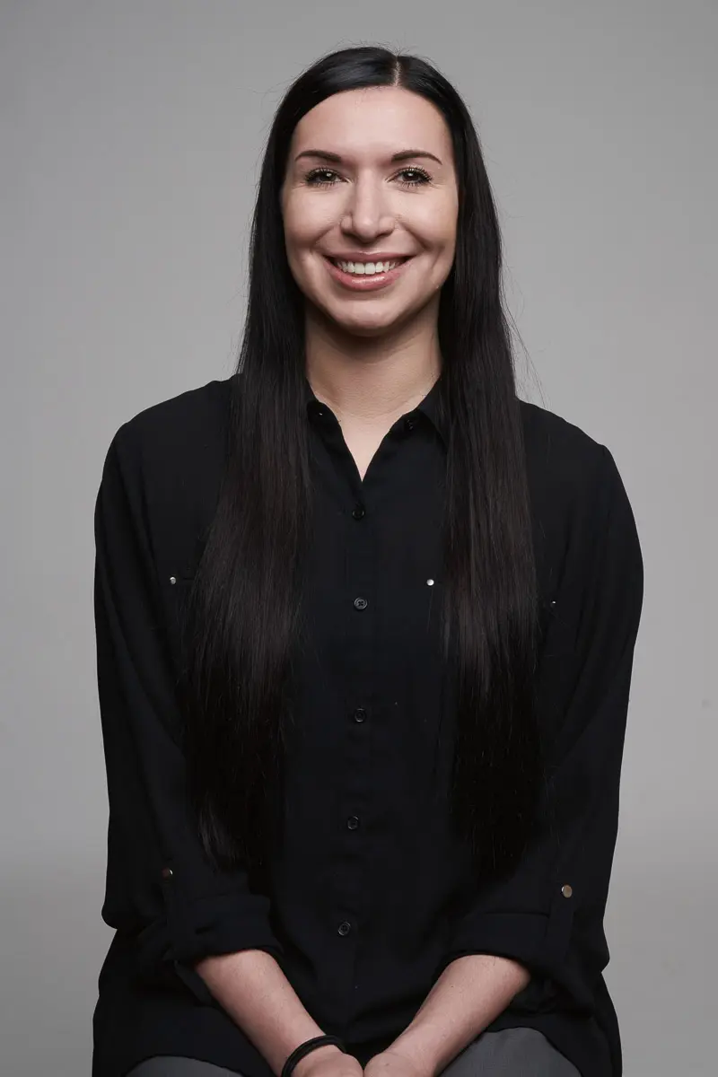 A woman with long straight dark hair, wearing a black button-up shirt, sits and smiles at the camera against a plain gray background.